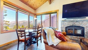 Ein gemütliches Wohnzimmer mit Kamin, großen Fenstern mit Blick auf die Berge, einem Essbereich mit Stühlen und einem an der Wand befestigten Flachbildfernseher.