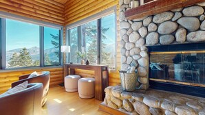 Interior de una cabaña de troncos con chimenea de piedra, paredes de madera y ventanas amplias con vistas a la montaña.