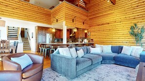 Interior acogedor de una cabaña con techo de madera, una sala de estar con sofá y sillones, una cocina con armarios de madera y una escalera que lleva a un nivel superior.