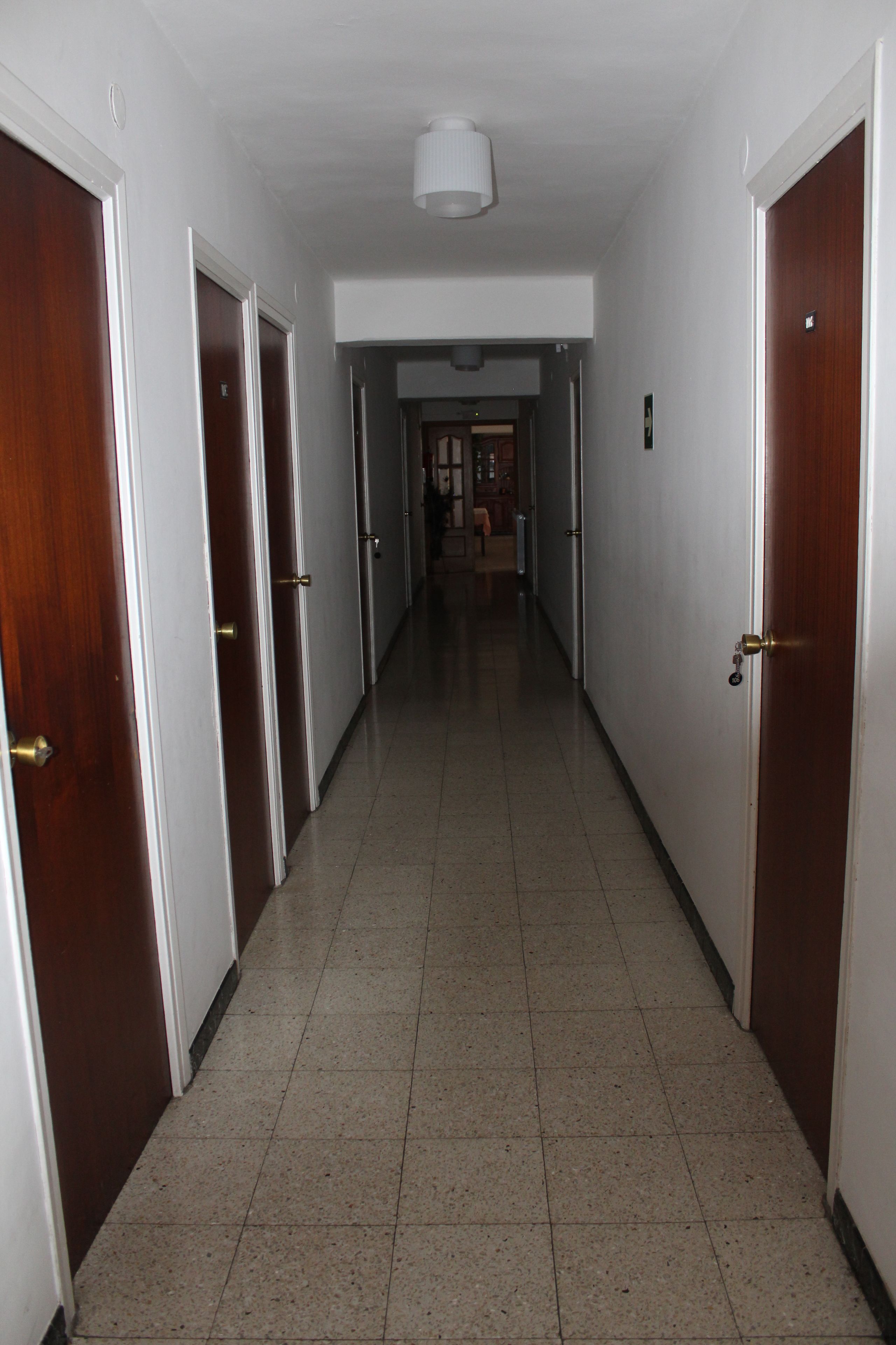 A corridor with wooden doors on both sides, leading to a room with a bed and a nightstand.