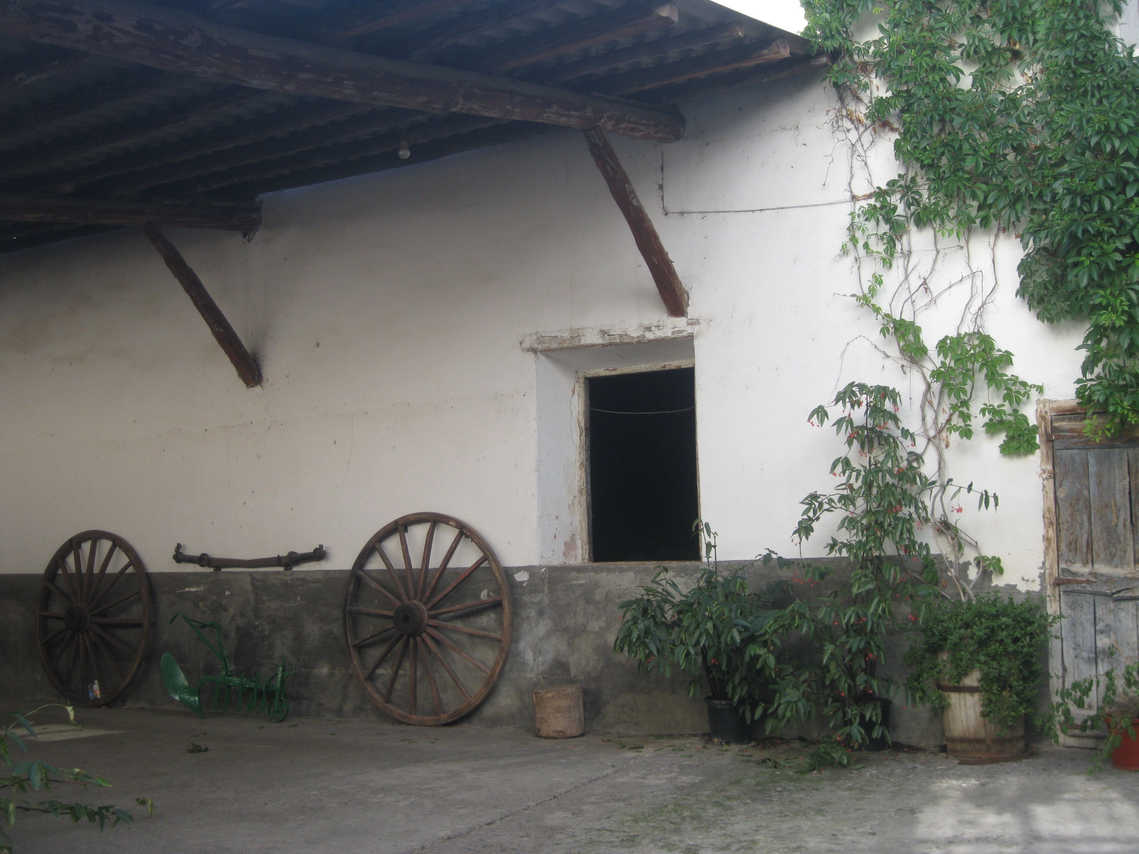 Un edificio rústico con fachada blanca, vigas de madera, una noria grande y plantas en macetas.