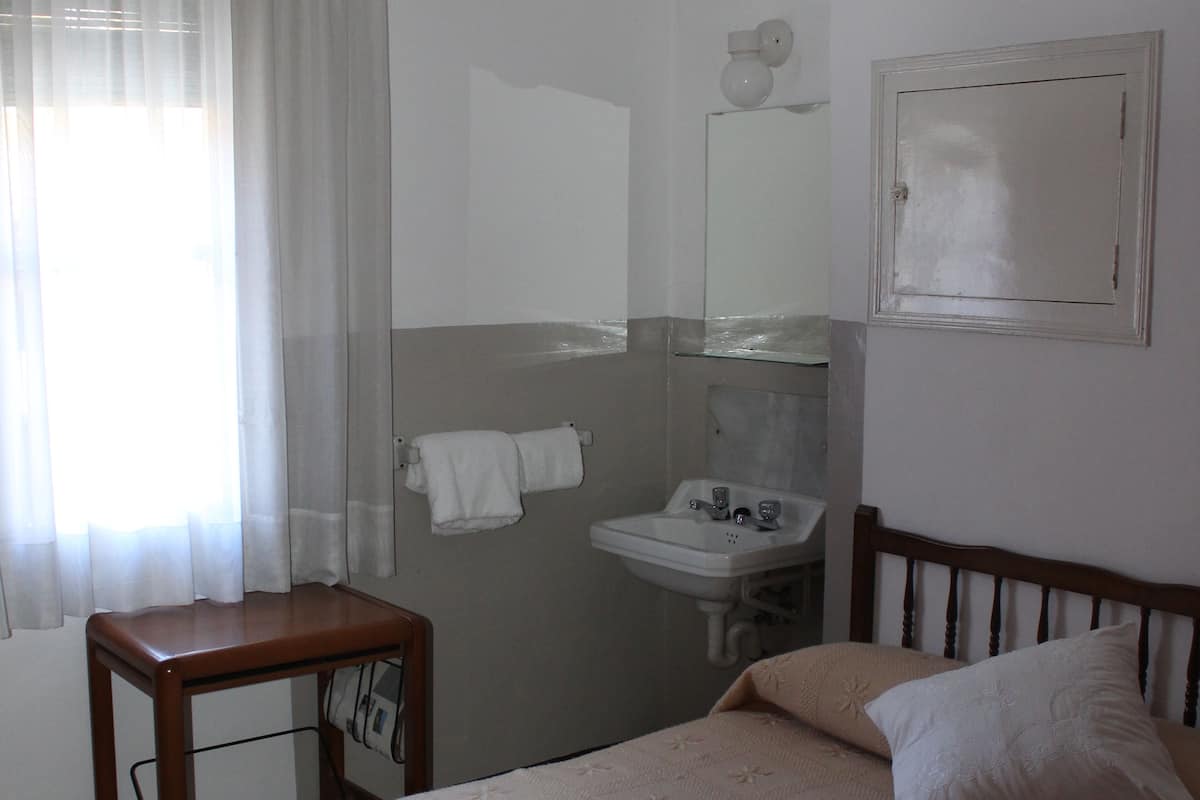 A bedroom with a bed, a bedside table, a window with curtains, a sink, and a mirror.