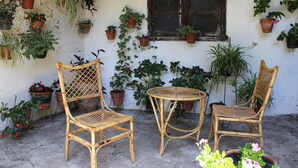 A room with two wicker chairs, a small round table, and various potted plants against a white wall with a window. - Pensión Peperillo (Graus)