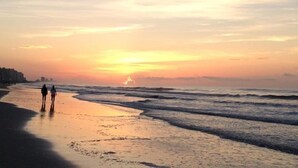 Una playa con olas al atardecer, cielo despejado y algunas personas caminando.