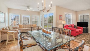 A spacious living room with a glass-top dining table, wicker furniture, a red sofa, and a chandelier. - The Cotton Patch in Cherry Grove: Walking Distance to Beach! Private Pool! (North Myrtle Beach)