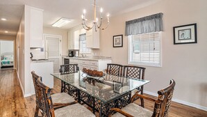 A dining area with a glass table surrounded by wicker chairs, a chandelier, and a kitchen in the background. - The Cotton Patch in Cherry Grove: Walking Distance to Beach! Private Pool! (North Myrtle Beach)