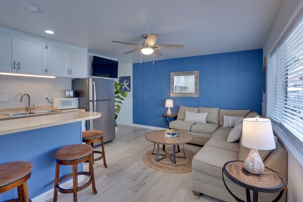 Eine moderne Küche mit weißen Schränken, einer Mikrowelle und einem Kühlschrank. Es gibt eine Essecke mit einem runden Holztisch und Hockern. Das Wohnzimmer verfügt über ein beiges Sofa und eine Lampe auf einem Beistelltisch.