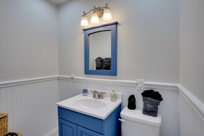 Ein Badezimmer mit einem weißen Waschbecken, einer blauen Kommode und einer Toilette. Über dem Spiegel befinden sich drei Leuchten, und eine Handtuchkorb hängt an der Wand.