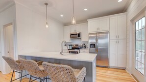 Una cocina moderna con armarios blancos, electrodomésticos de acero inoxidable, una isla central con taburetes y luminarias colgantes.