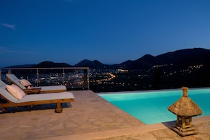 A poolside area with lounge chairs and a thatched hut, overlooking a cityscape and mountains at dusk. - Luxury villa overlooking the sea with private pool-Villa Dahlia (Lefkada)