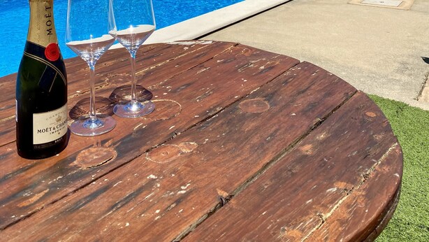 Ein Holztisch mit einer Champagnerflasche und zwei Gläsern, ein Swimmingpool und Liegestühle mit einem weißen Sonnenschirm.