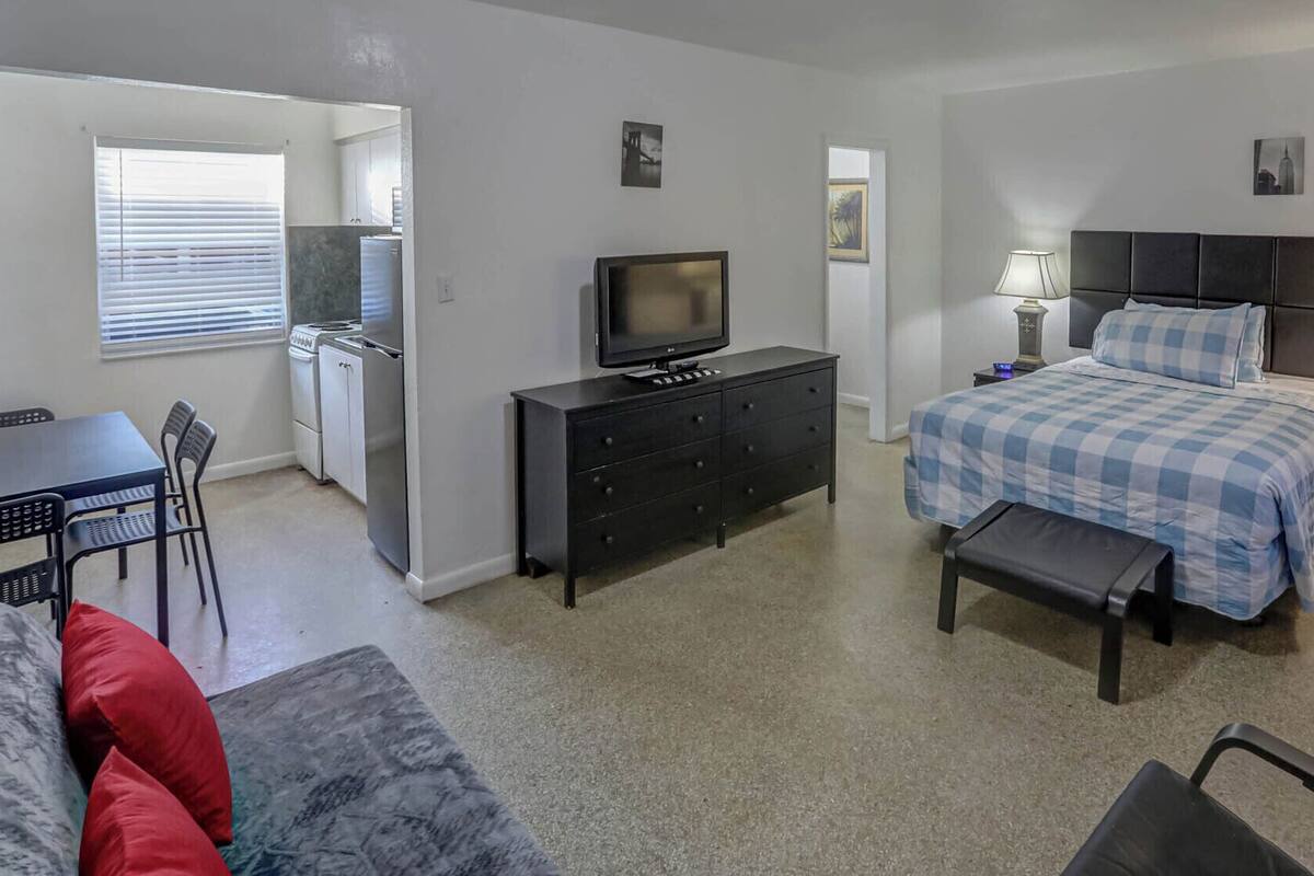 A compact hotel room with a bed, a desk, a chair, a television, and a small kitchenette.