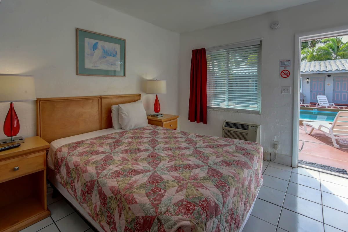 A hotel room with a large bed, two bedside tables, a nightstand, a window with blinds, a red curtain, and a view of an outdoor pool area.