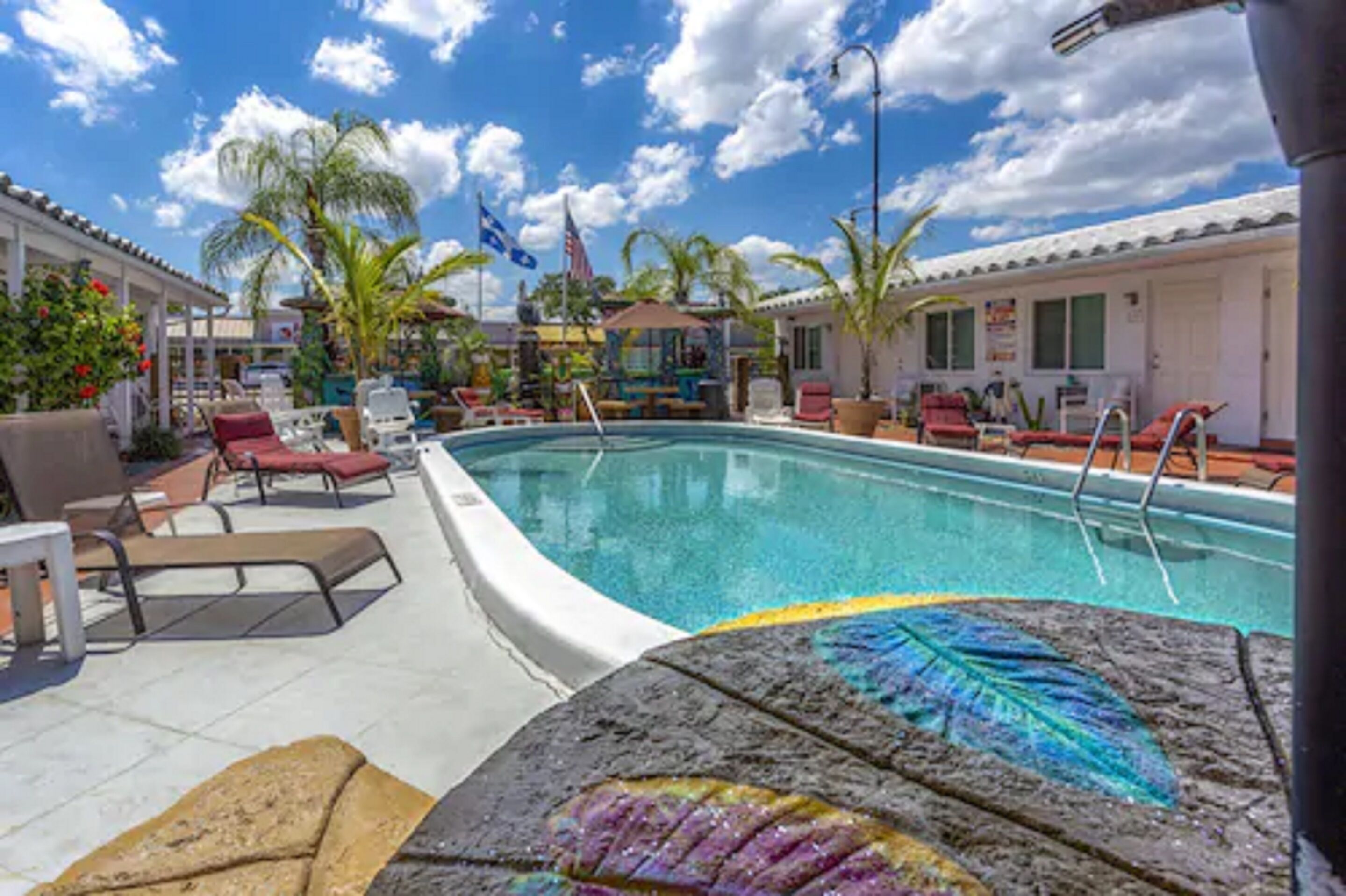 A pool area with lounge chairs, a flag, and a building with a tiled roof.