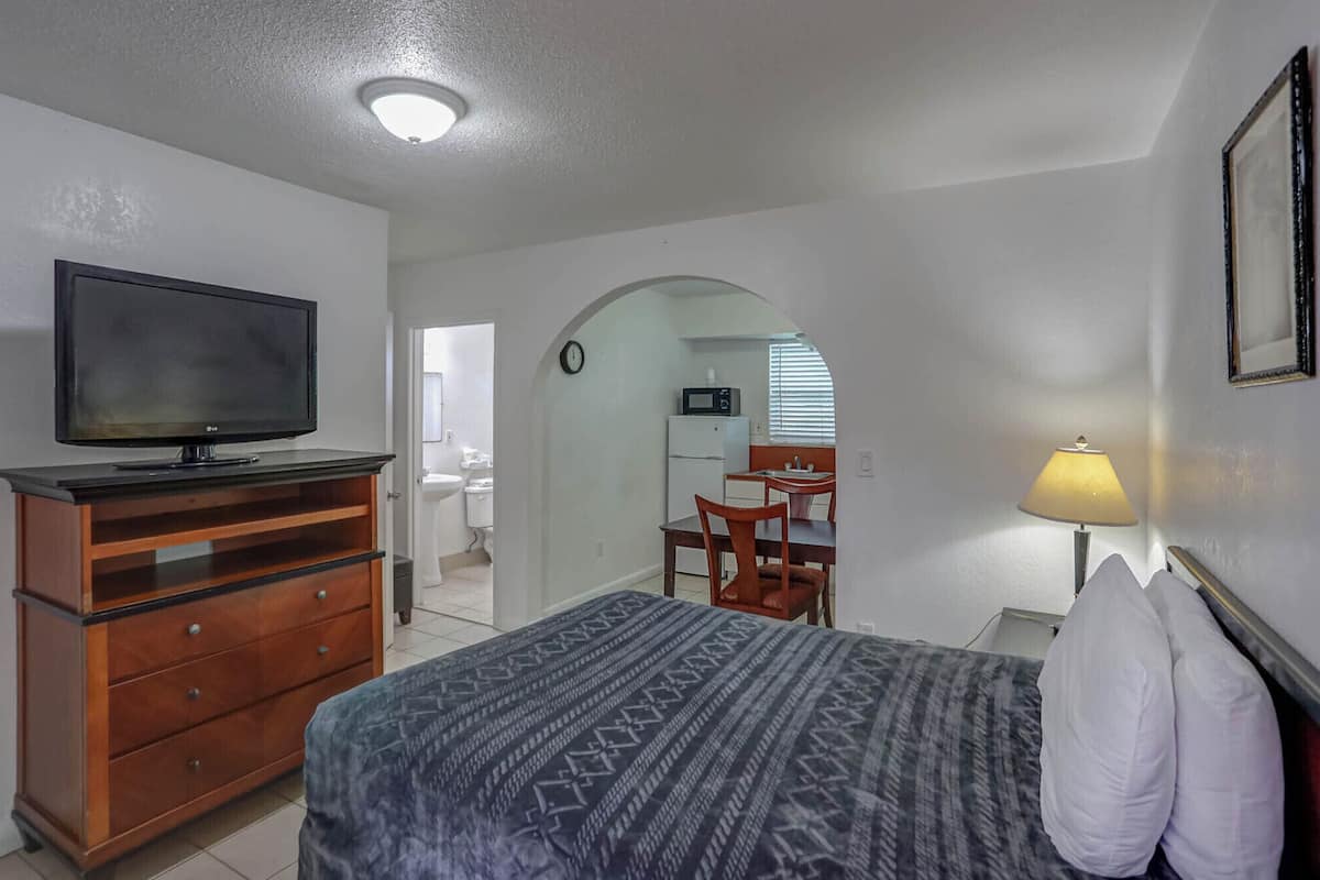 A bedroom with a bed, dresser, TV, lamp, and an archway leading to a bathroom and kitchen.