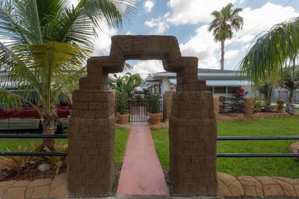 A pathway leading through a stone archway, flanked by palm trees and a building in the background.