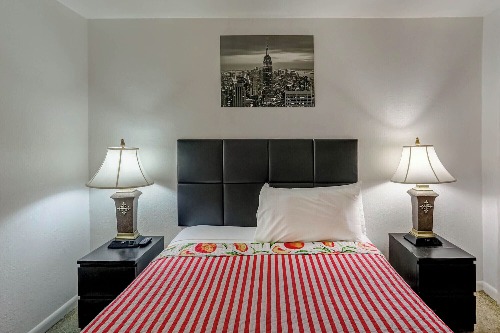 A hotel room with a bed, two bedside tables, two lamps, a black headboard, a white pillow, a red and white striped bedspread, and a cityscape painting on the wall.