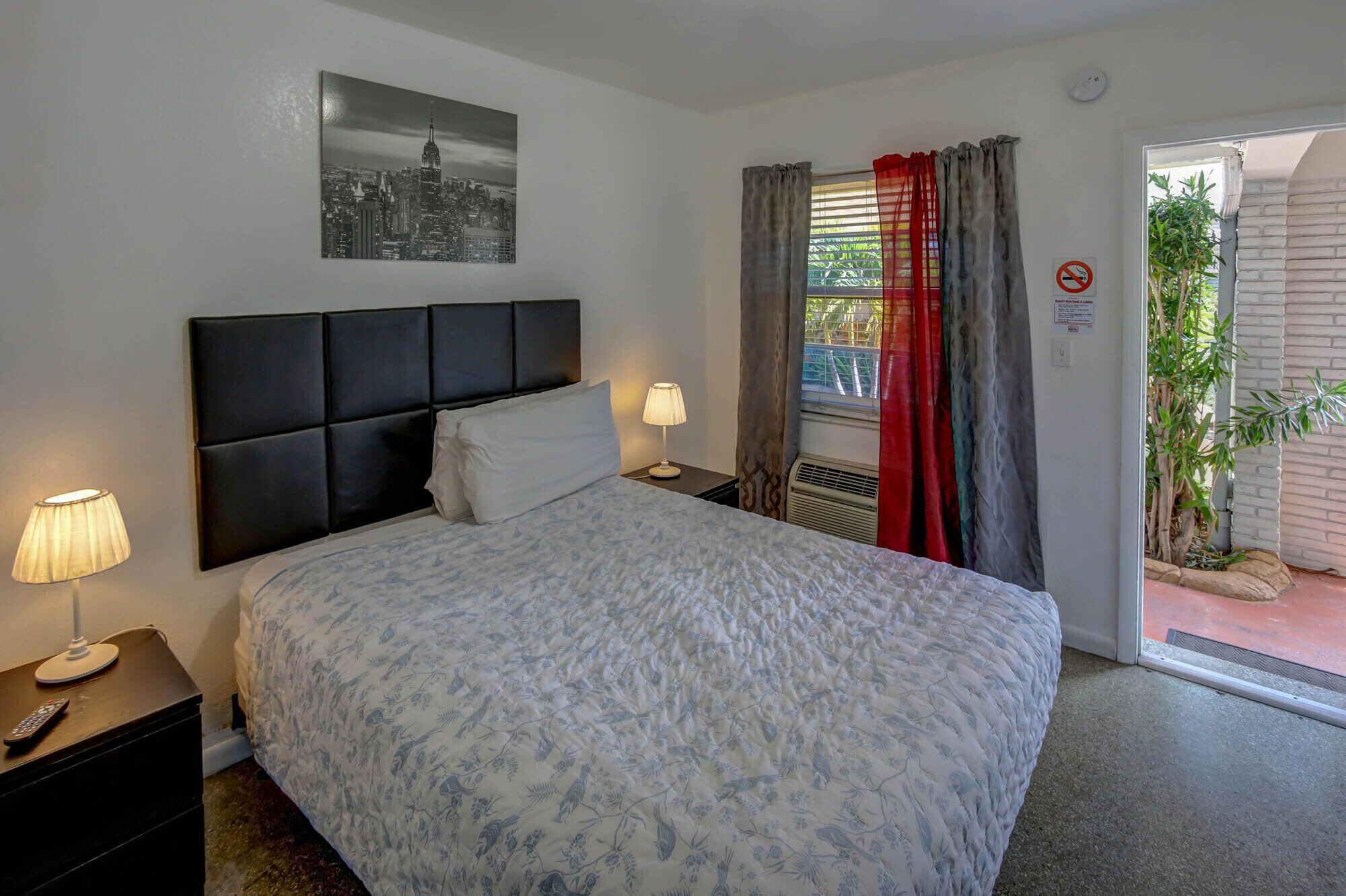 Ein Hotelzimmer mit einem Bett, einem Nachttisch, einer Lampe, einem Fenster mit Vorhängen und einem Stadtbild an der Wand.