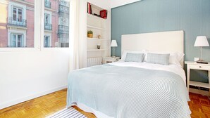 Un dormitorio con una cama grande, una mesita de noche, una lámpara, una estantería y vistas a un edificio.