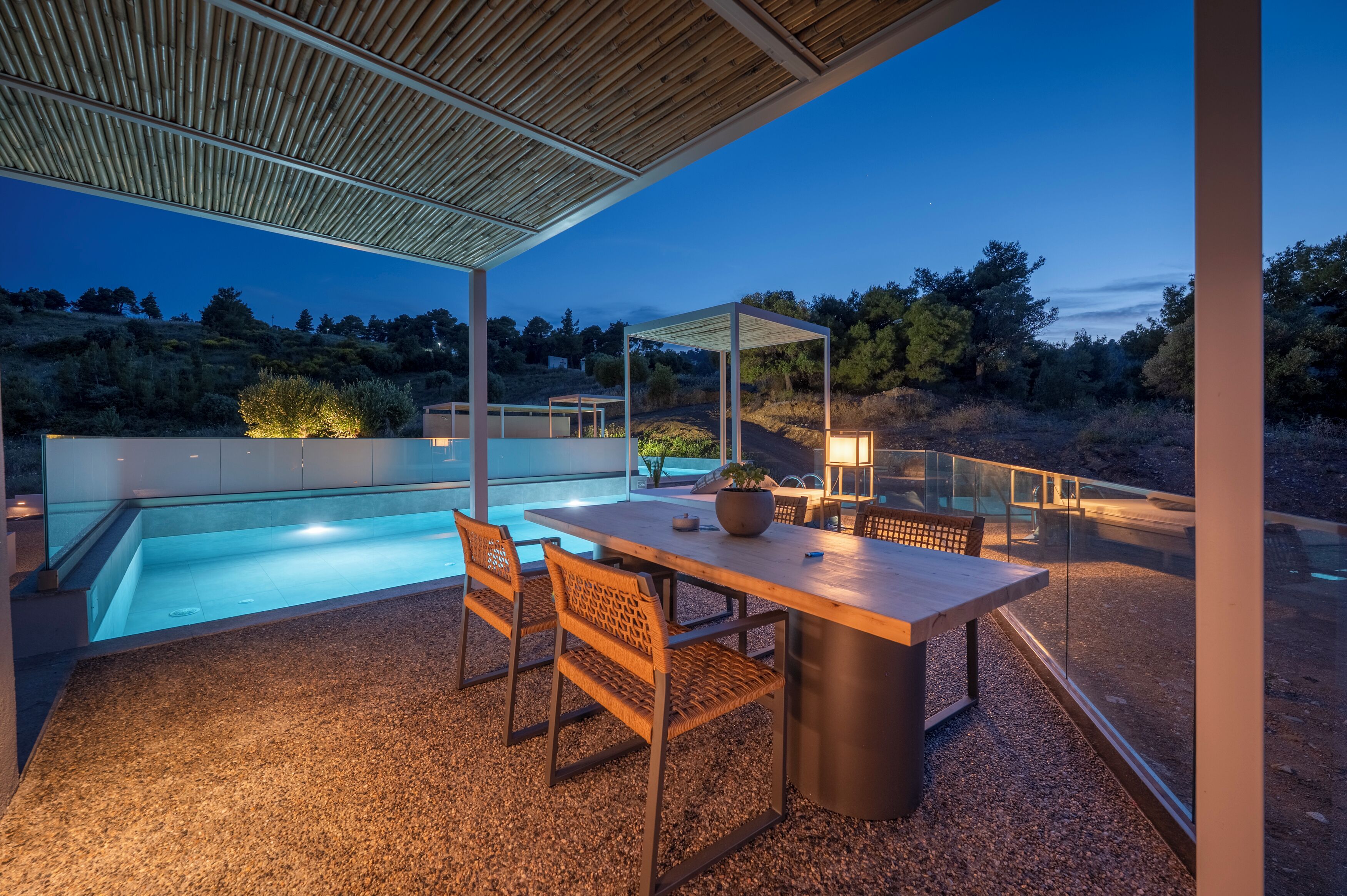 Un área de piscina exterior moderna con una mesa y sillas de madera, una pérgola y vistas del paisaje circundante.