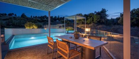 Un área de piscina exterior moderna con una mesa y sillas de madera, una pérgola y vistas del paisaje circundante.