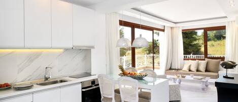 A modern kitchen with white cabinetry, a marble backsplash, and a central island with chairs. There is a dining area with a glass table and chairs, and a sofa with a view of the outdoors.