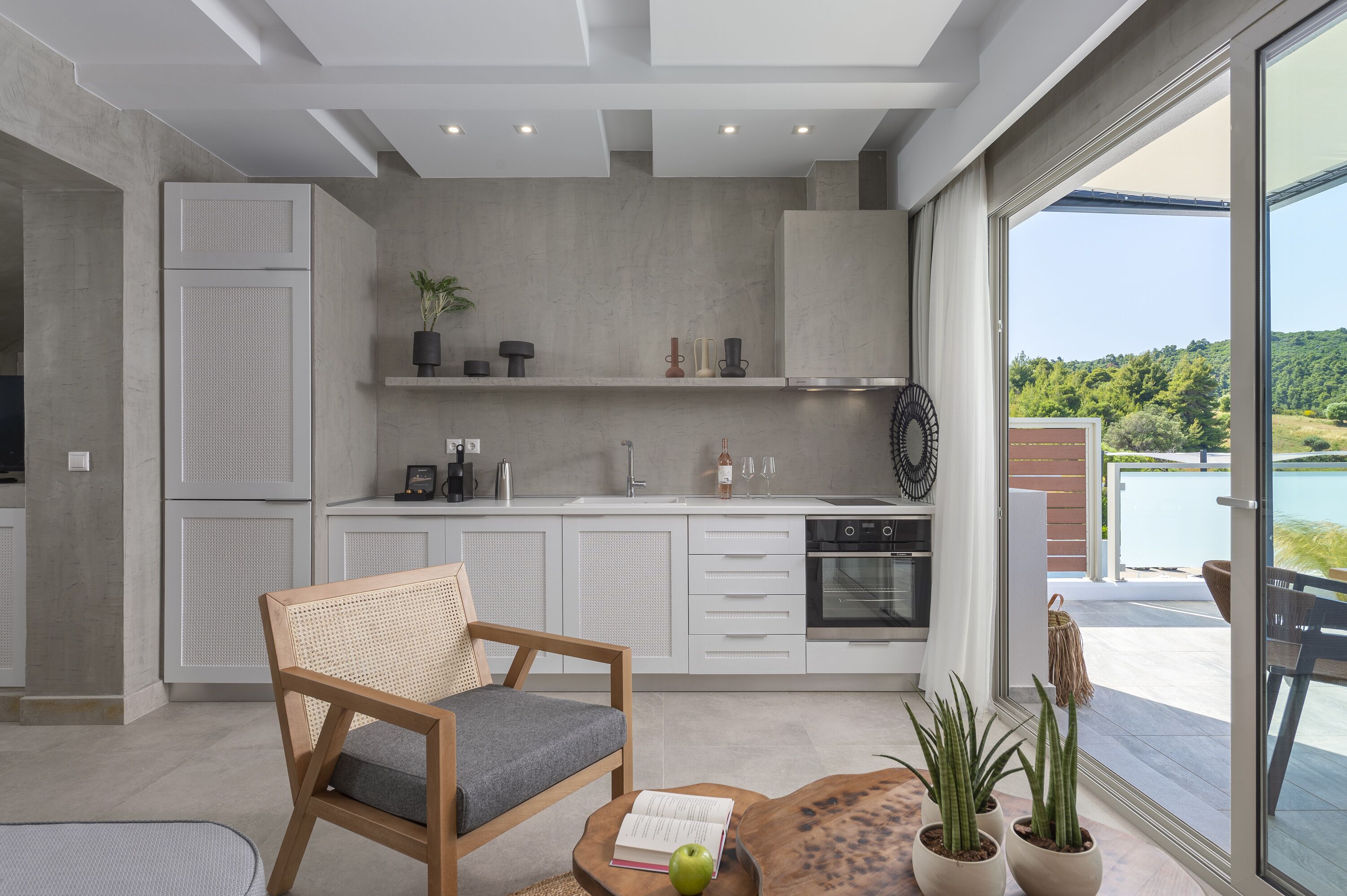 A modern kitchen with white cabinets, a dining area with a wooden table and chair, and a view of a lush landscape through large windows.