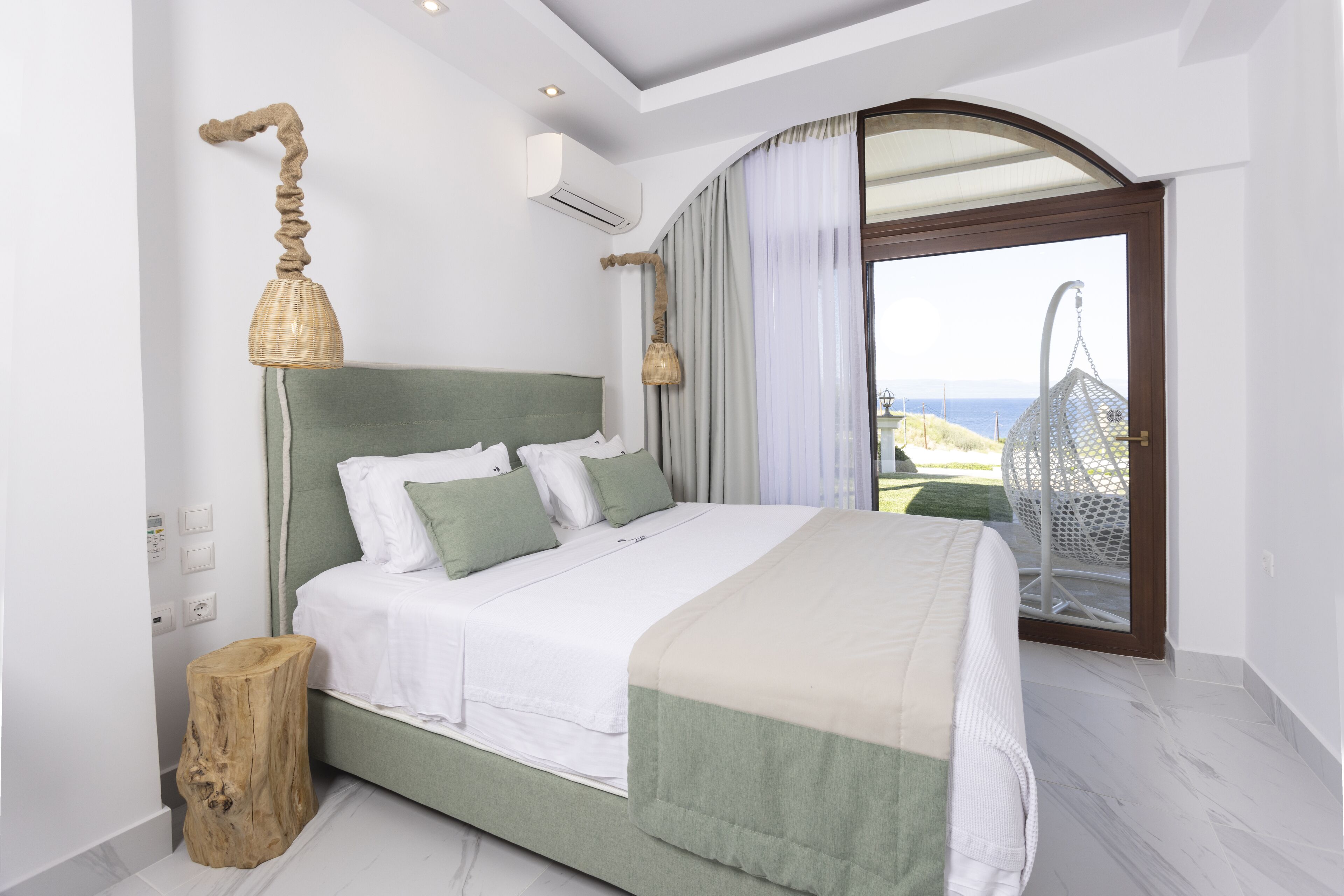 A bedroom with a bed, a nightstand, a lamp, a wooden stool, and a view of the sea through a large window.