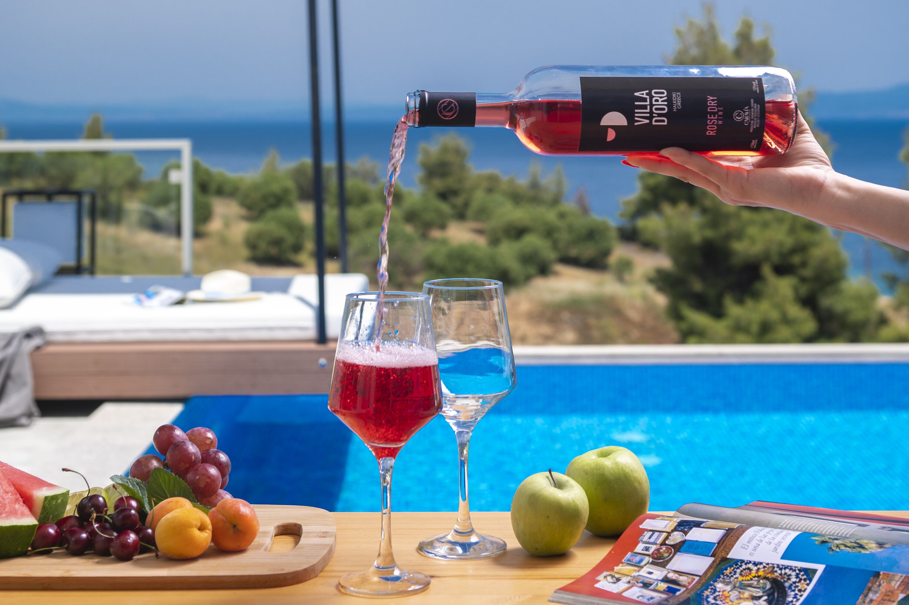 A hand pouring red wine into a glass by a poolside table with fruits and a magazine.