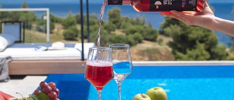 A hand pouring red wine into a glass by a poolside table with fruits and a magazine.