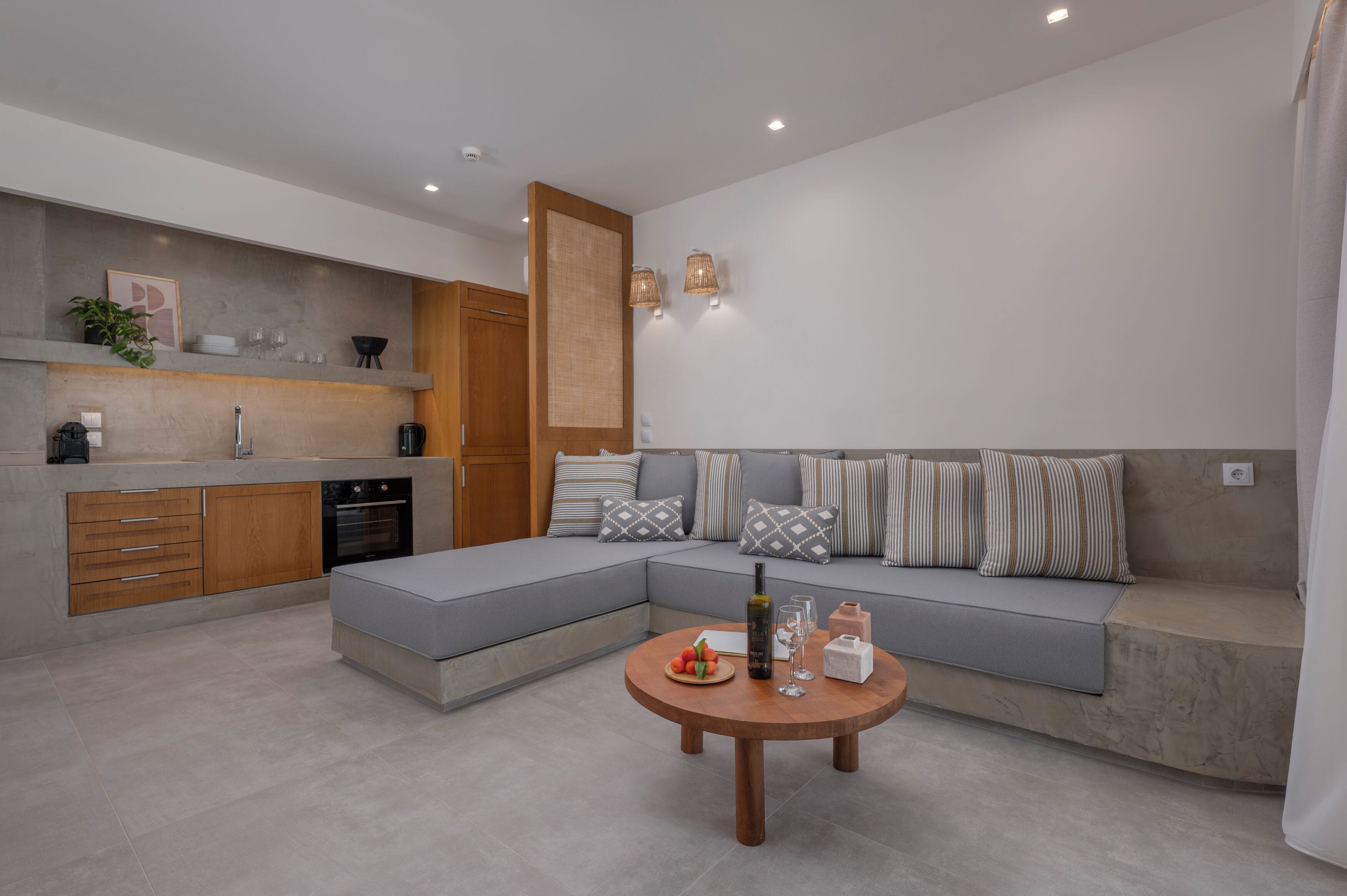 A modern kitchen with built-in appliances, a grey sectional sofa with patterned cushions, and a round wooden coffee table with a bottle and glasses.
