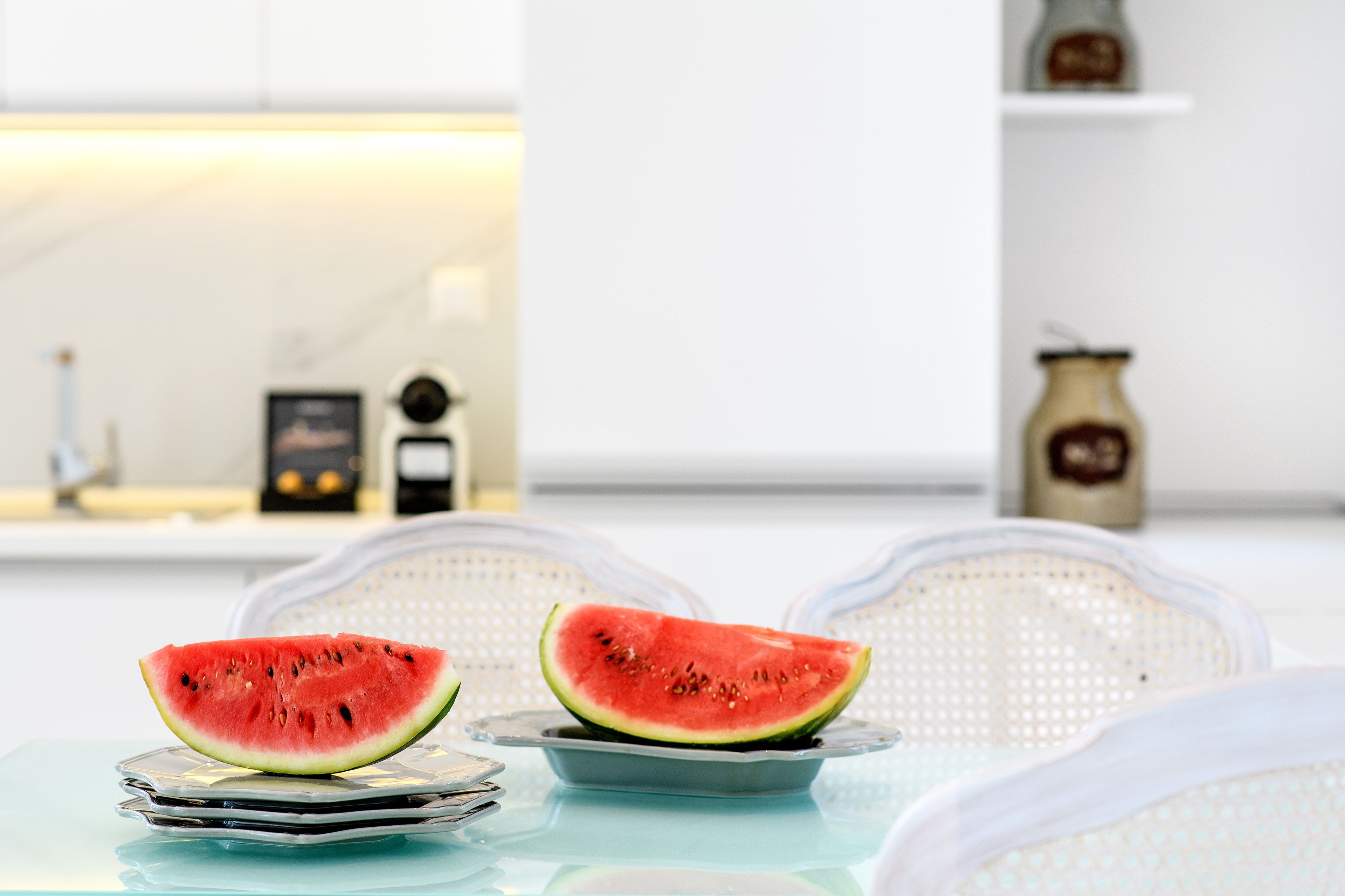 A table with two plates of watermelon slices, a jar, and a small clock on a white chair in a modern kitchen setting.