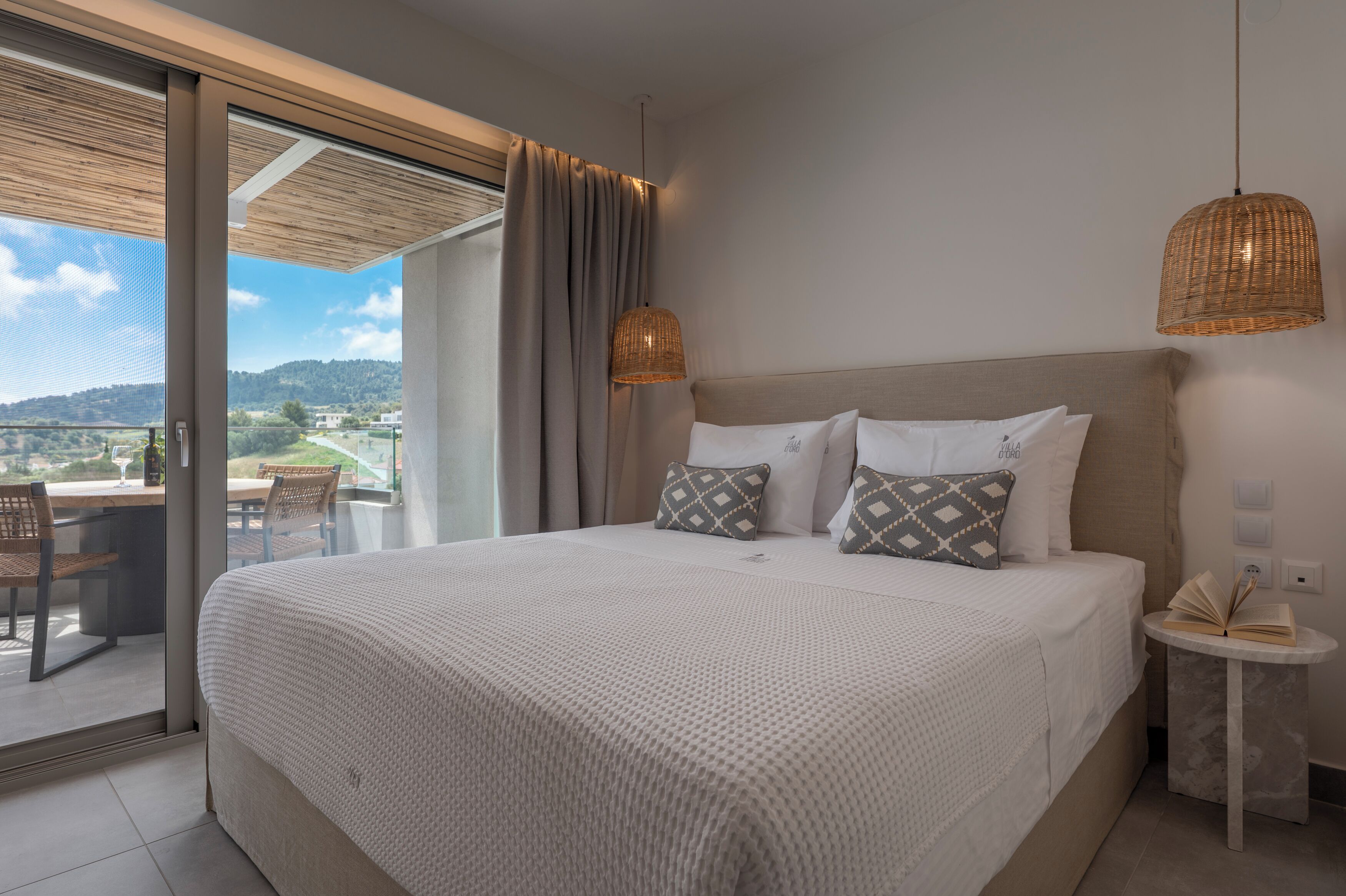 Un dormitorio con una cama grande, una mesita de noche pequeña, vista a través de una puerta corrediza de un espacio exterior con sillas, y una lámpara colgante de mimbre.