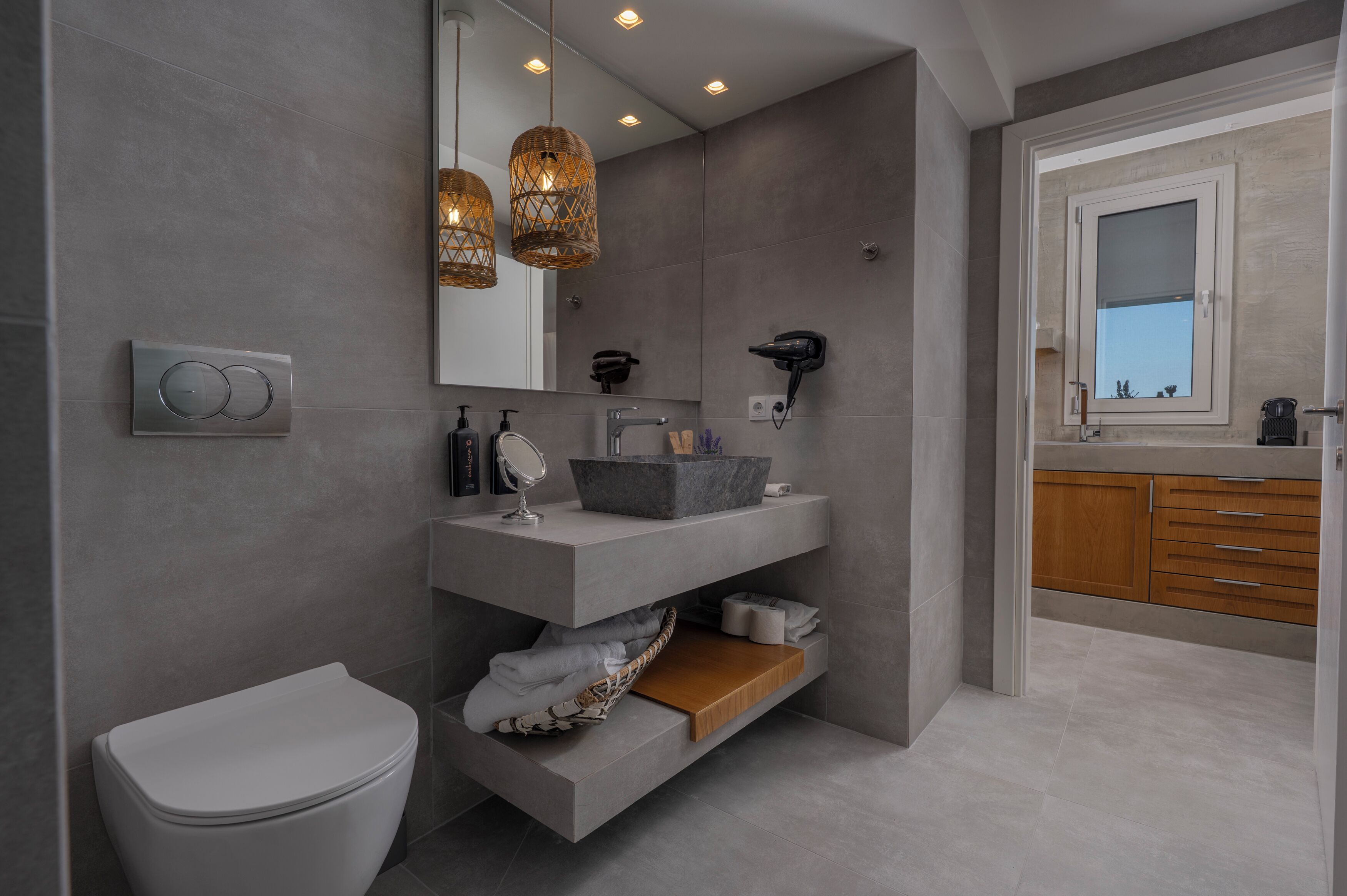 A modern bathroom with a grey tiled wall, a white toilet, a grey vanity with a marble basin, a wooden shelf, and a window with a view.