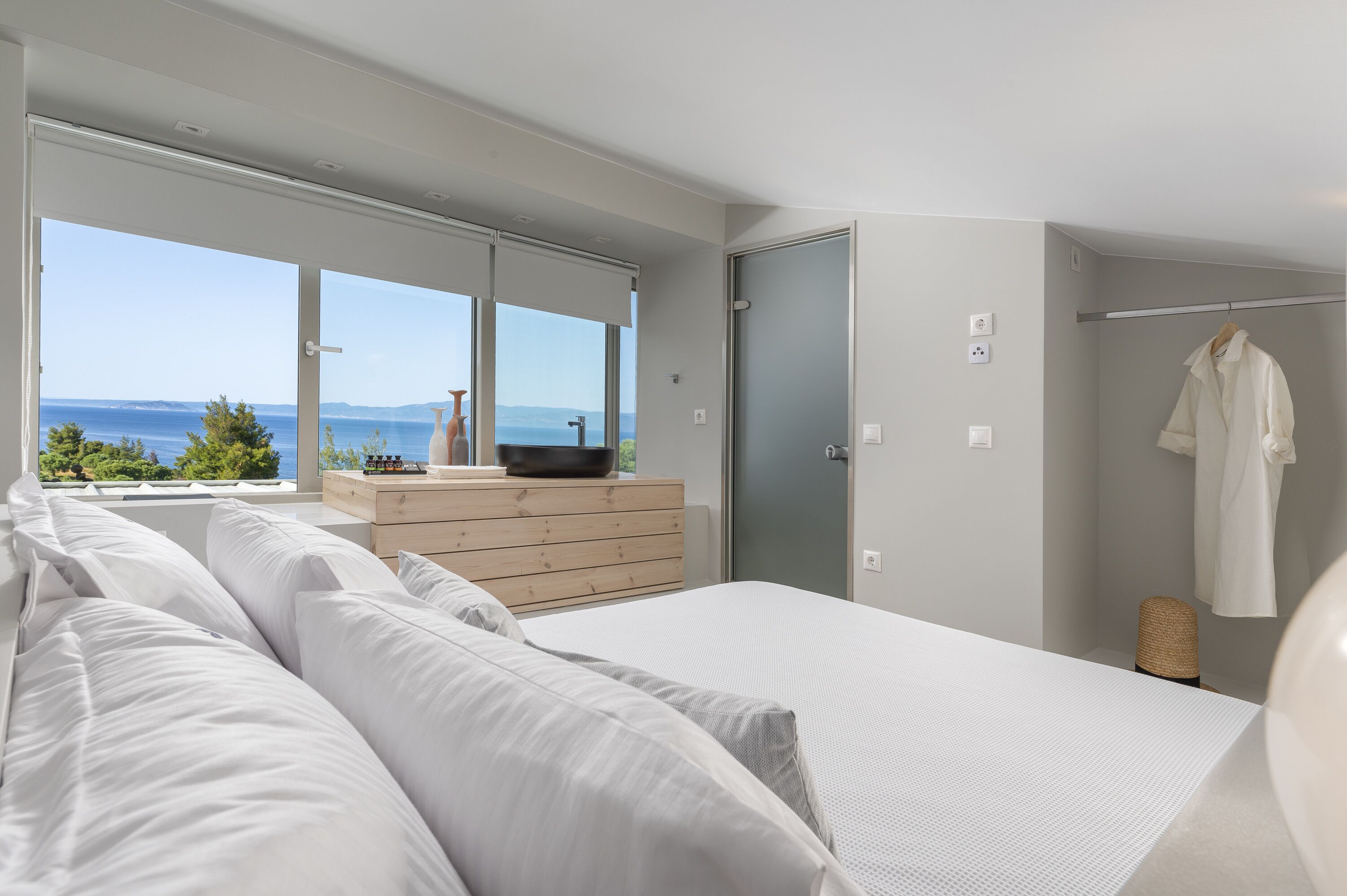 A modern bedroom with a large bed, a wooden dresser, and a view of the sea.