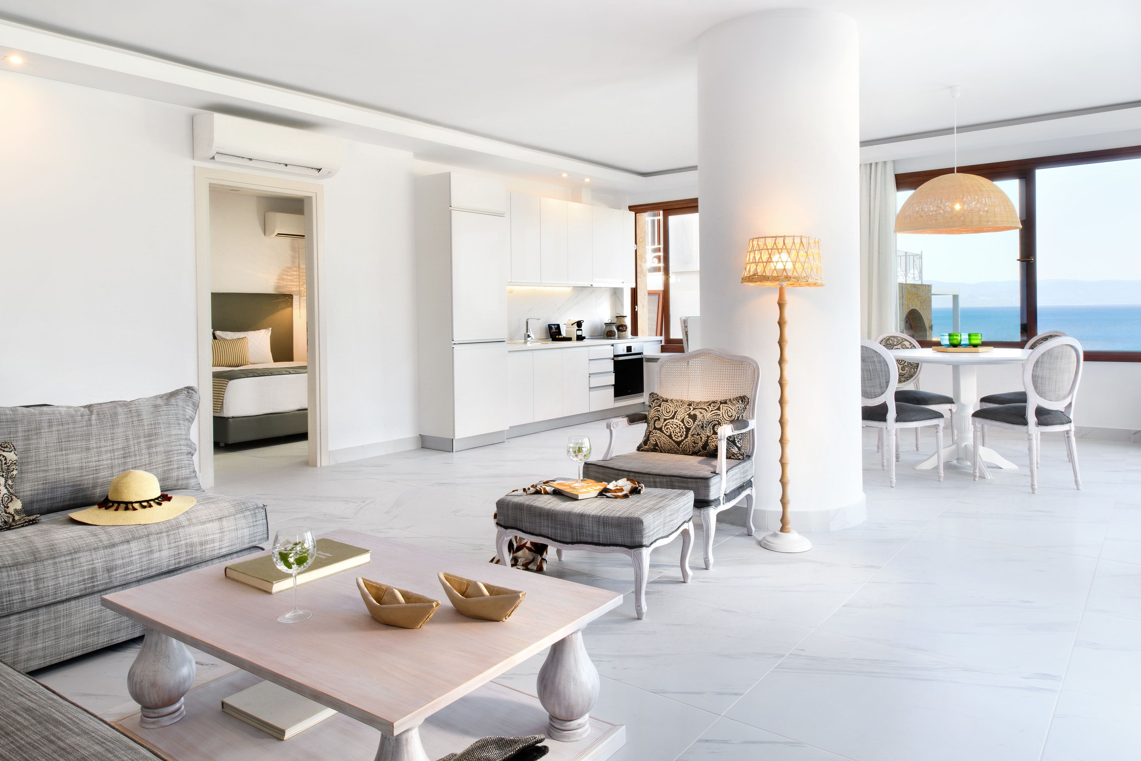A modern living room with a sofa, coffee table, and ottoman, a dining area with chairs and a table, a kitchen with white cabinets, and a large window with a view.