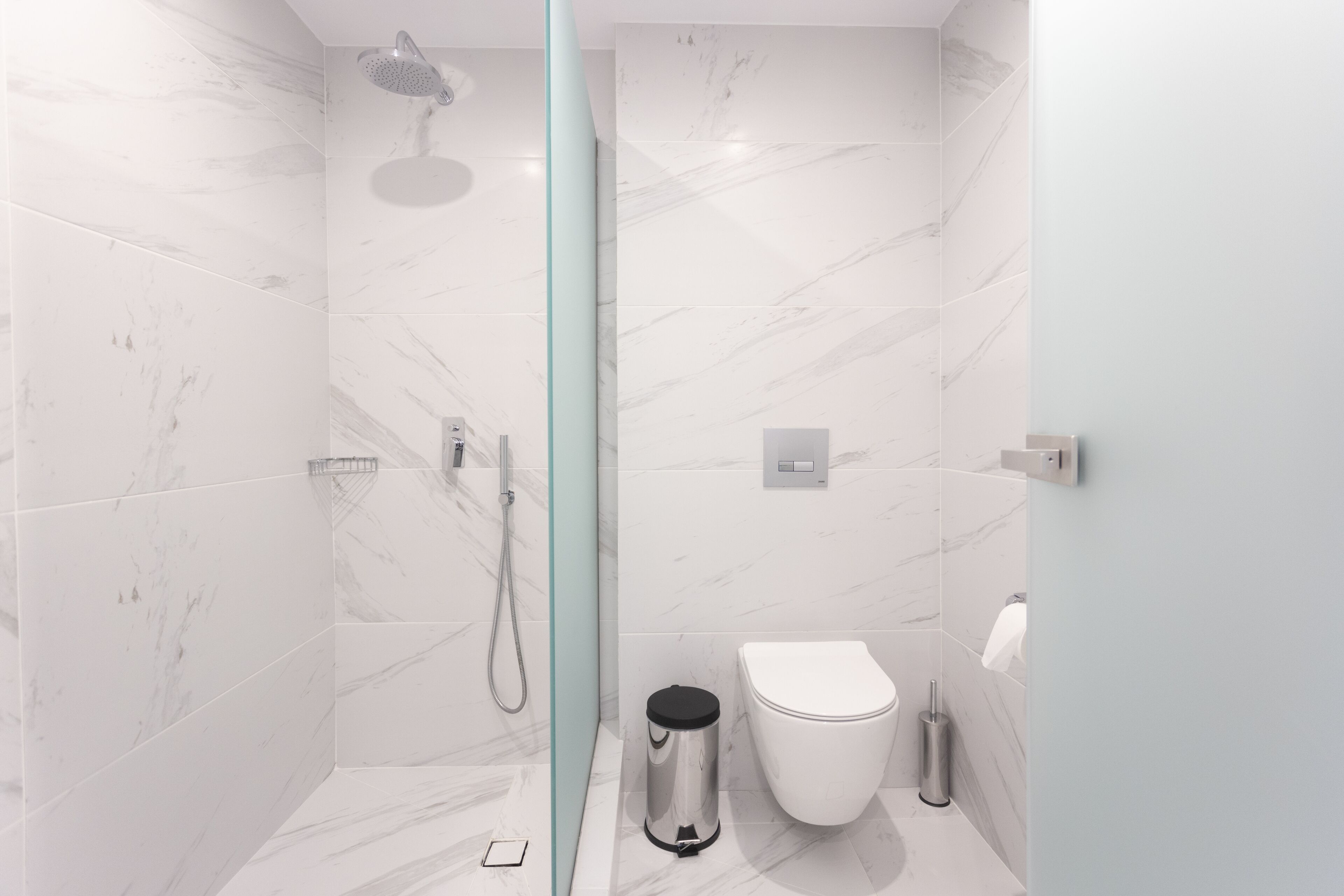 Een moderne badkamer met een douchecabine van glas, een wit toilet en een roestvrijstalen prullenbak.