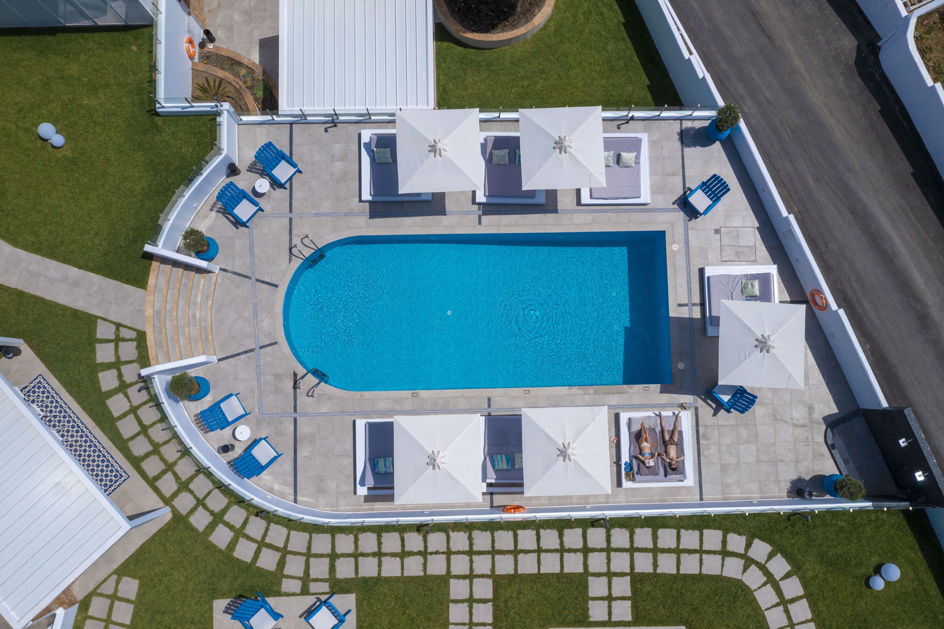 An aerial view of a swimming pool area with lounge chairs and umbrellas, surrounded by a paved deck and a small garden.