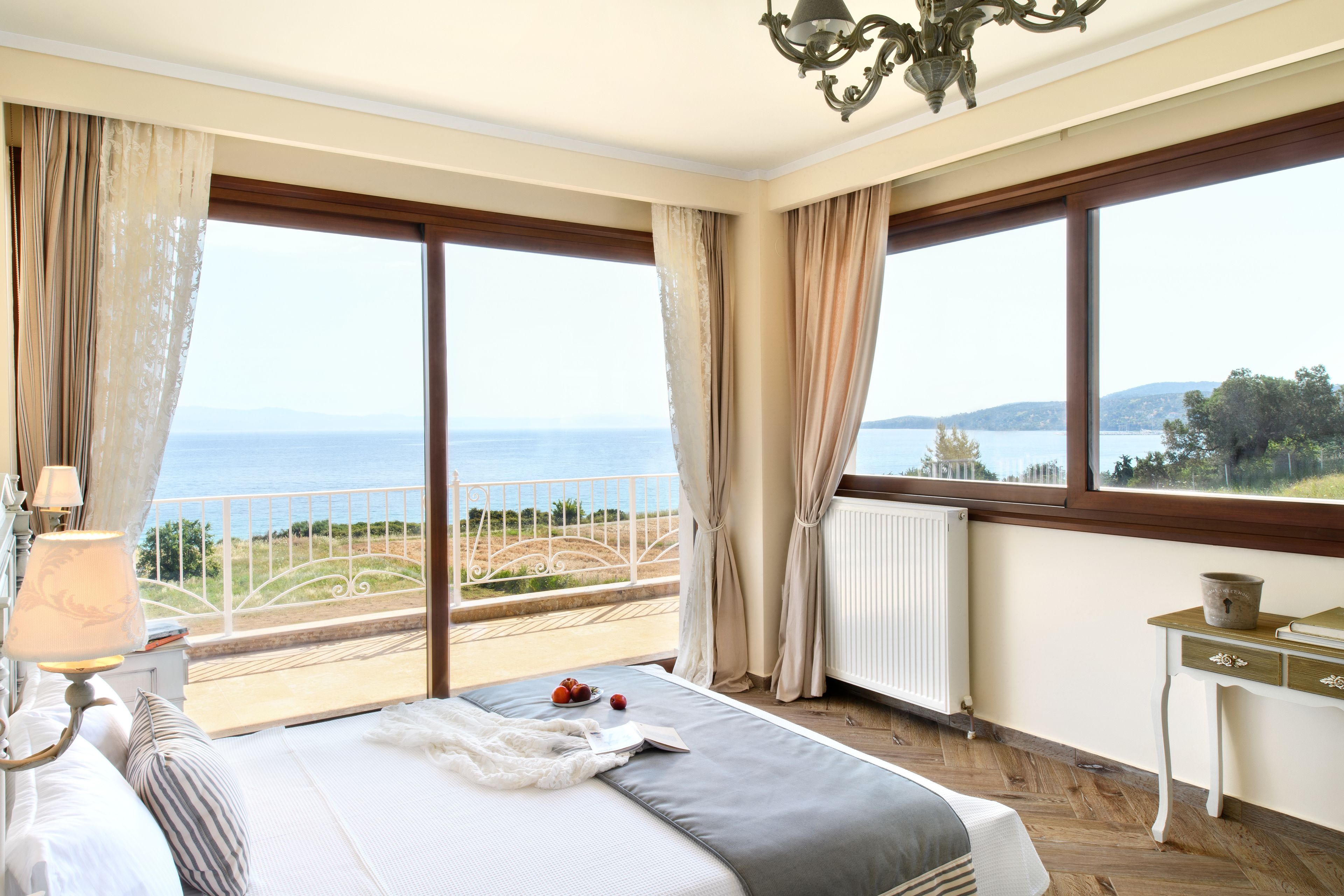 A bedroom with a large bed, a bedside table with a lamp, a view of the sea, and a window with curtains.
