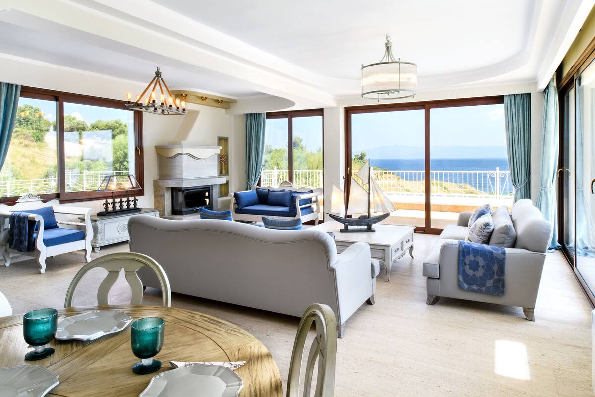 Uma sala de estar ampla com lareira, assentos confortáveis e grandes janelas com vista para o mar.