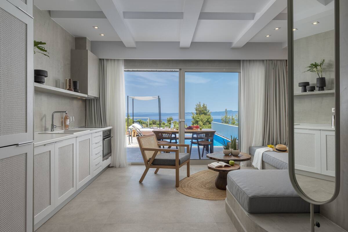 Cozinha moderna com armários brancos, área de jantar com mesa e cadeiras, sofá e vista para o mar através de grandes janelas.