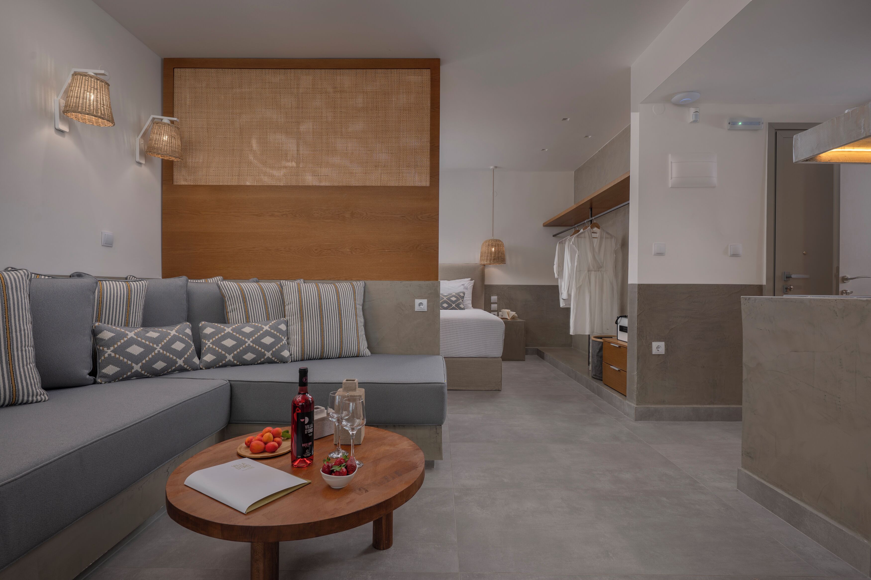 Een moderne hotelkamer met een bank, een eettafel met fruit en een fles, een bed met nachtkastje, en een badkamer met douche.