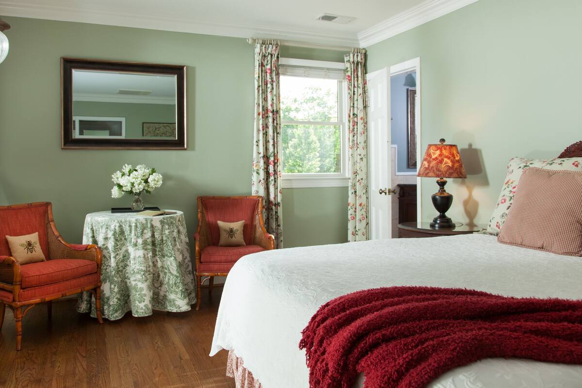Un dormitorio con una cama, dos sillones, una mesita con un florero y un espejo, además de una ventana con cortinas.