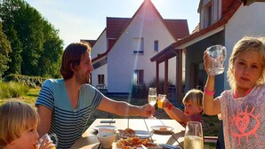 A family dining outdoors at a table with food and drinks, with a house in the background. - Terrace Stay near Shoreline (Berck-Sur-Mer)