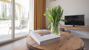 A modern living room with a wooden table, a glass vase with green plants, a stack of magazines, a TV on a wooden stand, and a sliding glass door leading to an outdoor area. - Terrace Stay near Shoreline (Berck-Sur-Mer)