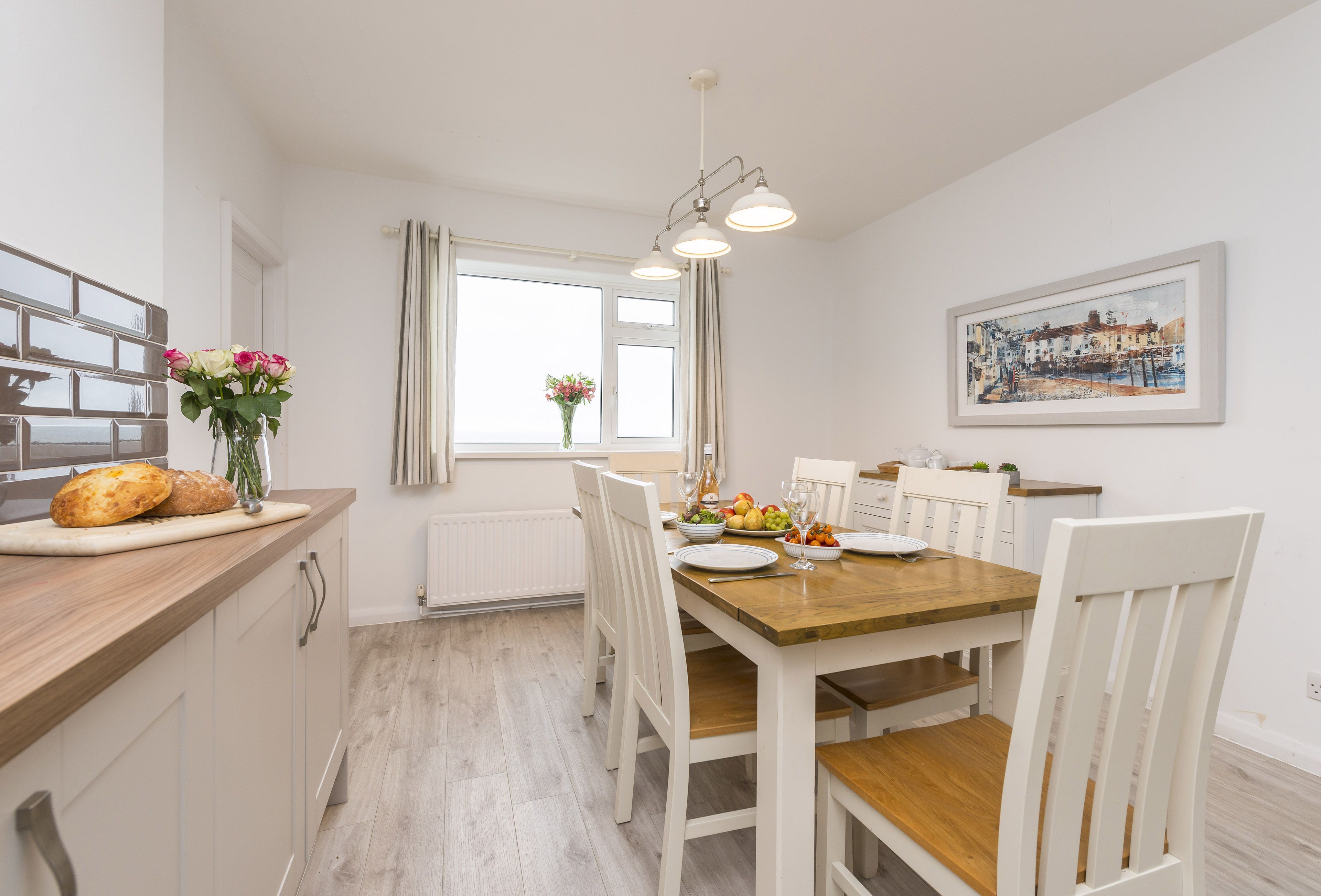 A dining area with a wooden table, white chairs, a window with curtains, a radiator, and a painting on the wall.