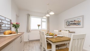 Een eetgedeelte met een houten tafel, witte stoelen, een raam met gordijnen, een radiator en een schilderij aan de muur.