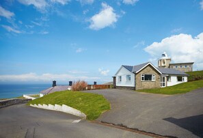 A coastal property with a stone house, a white building with a tower, and a paved driveway leading to a white fence.