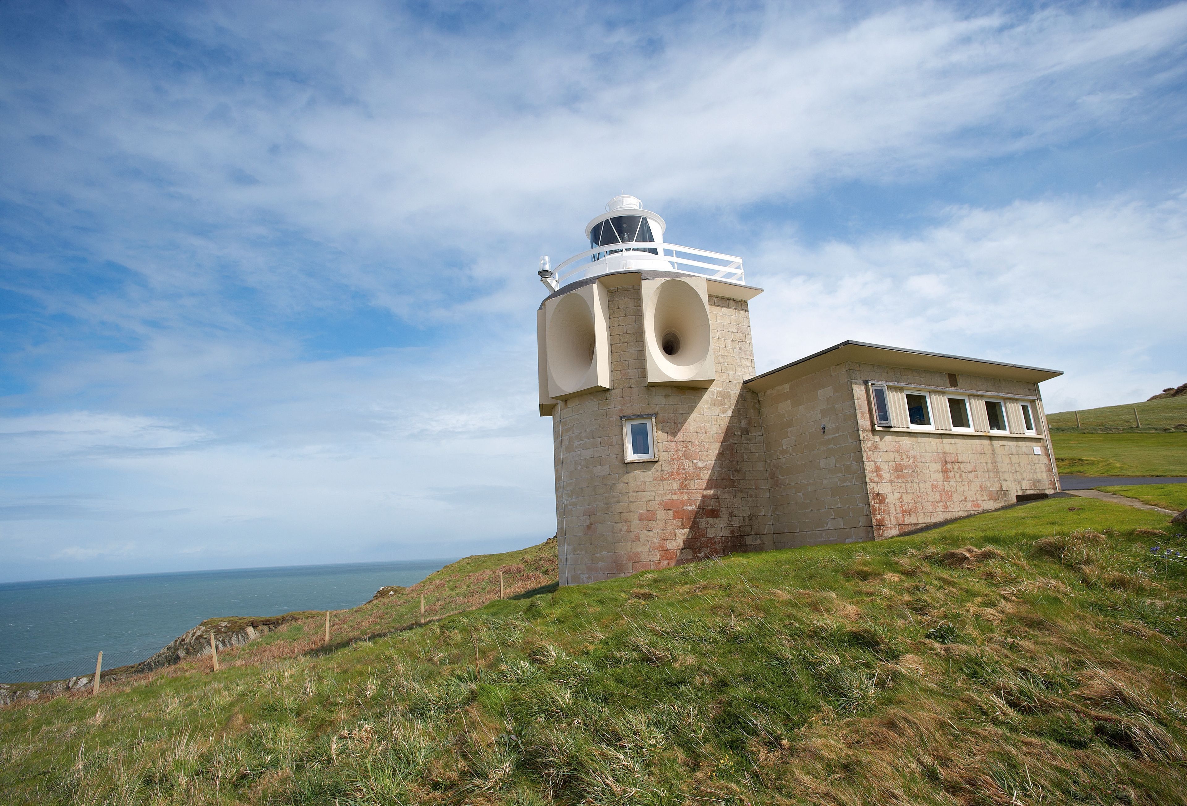 Ein Leuchtturm mit einem zylindrischen Turm und einem kleineren angrenzenden Gebäude, gelegen auf einer grasbewachsenen Anhöhe mit Blick aufs Meer.