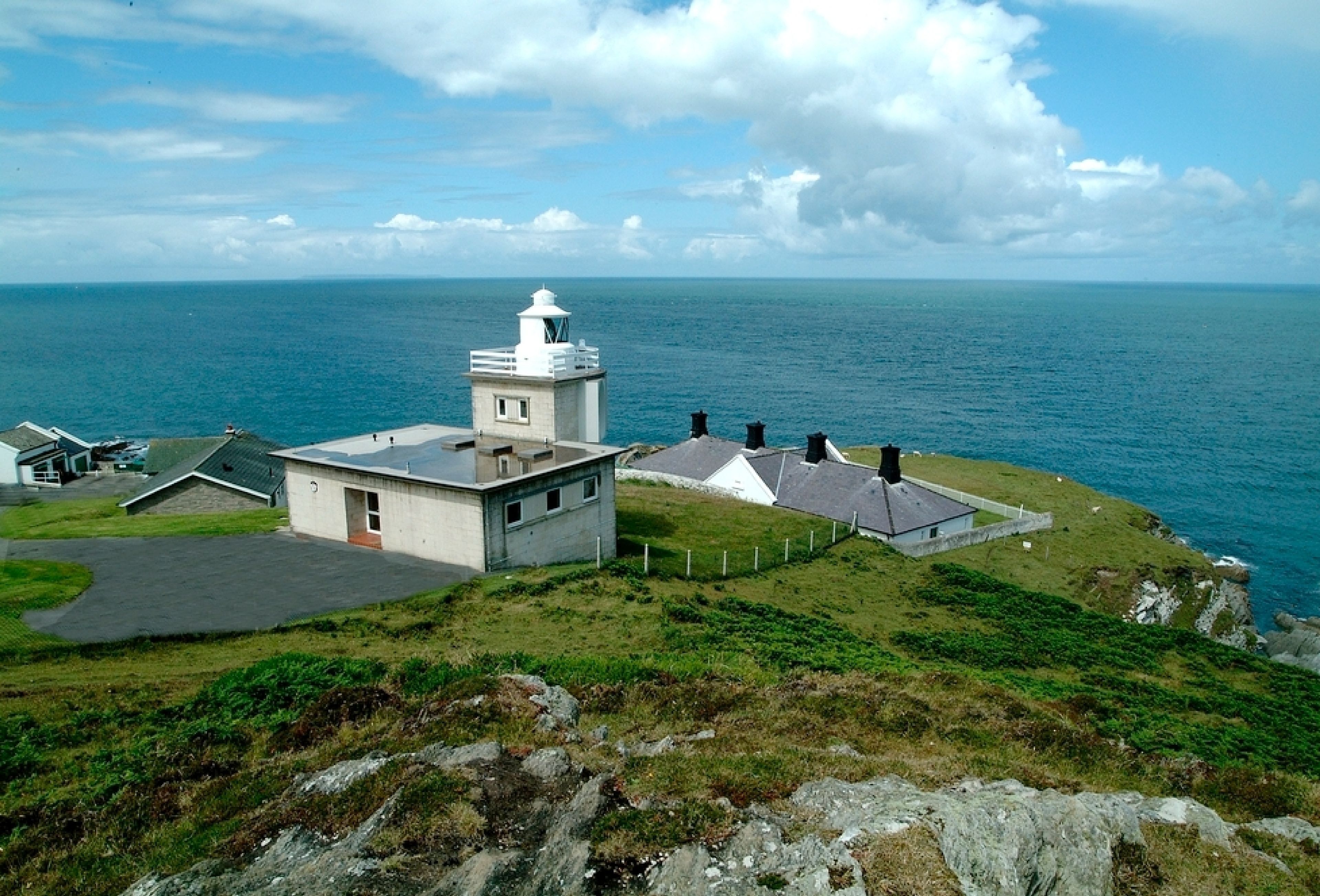 Um farol costeiro com torre branca e um pequeno edifício anexo, cercado por área gramada e mar.